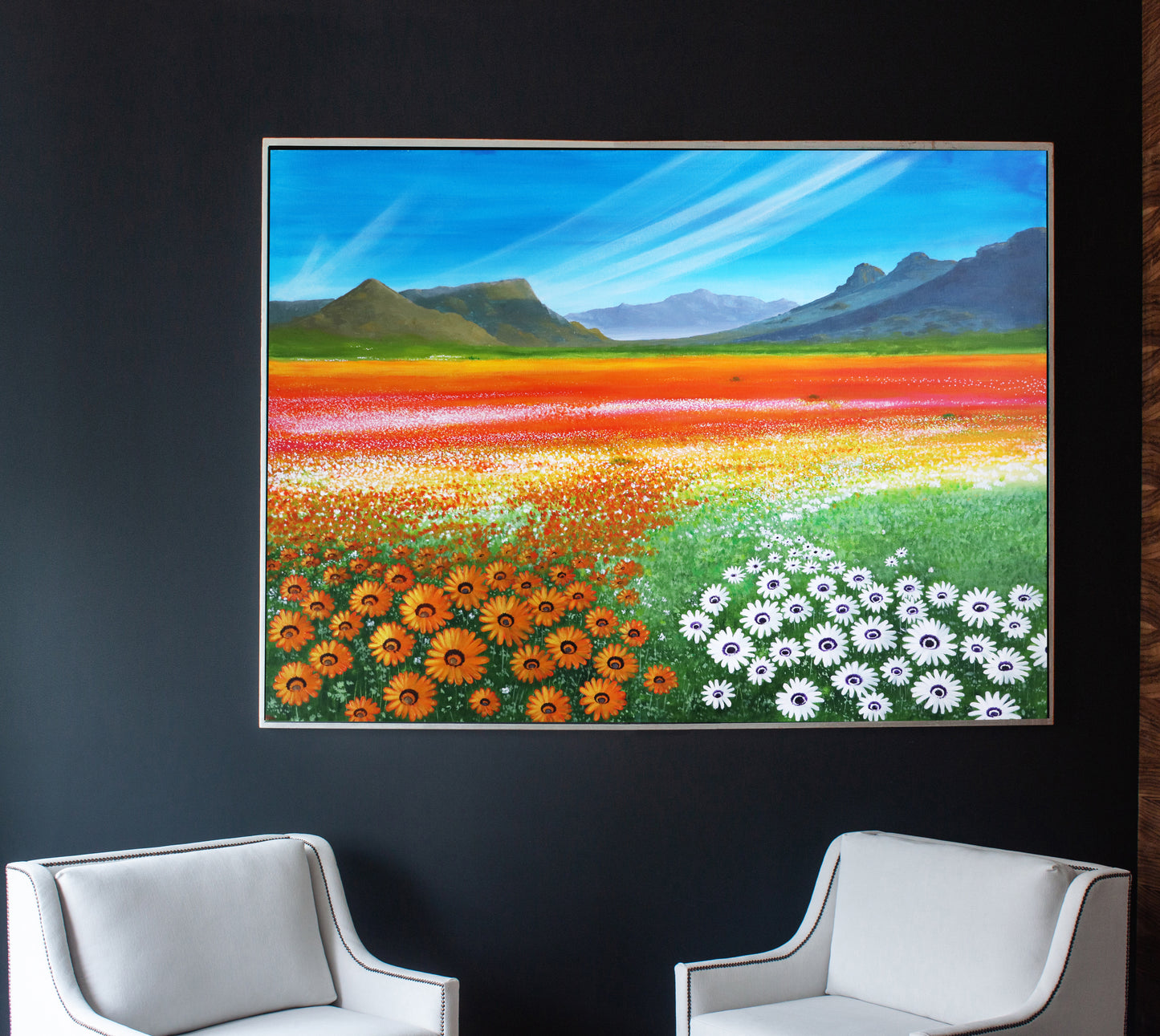 A vibrant painting of a landscape with fields of wild flowers in shades of orange, red, yellow, and white, with a blue sky and mountains in the background, displayed on a wall between two white chairs.